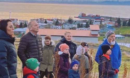 A group of adults and children in winter clothing stand on a hillside overlooking Húsavík, Iceland, with the town's coastal buildings and snowy mountains visible in the sunset background.