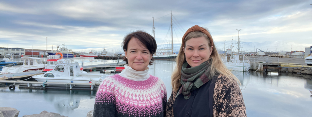 Two women, the co-founders of STEM Húsavík, stand smiling at the Húsavík harbor with fishing boats and snow-capped mountains in the background.