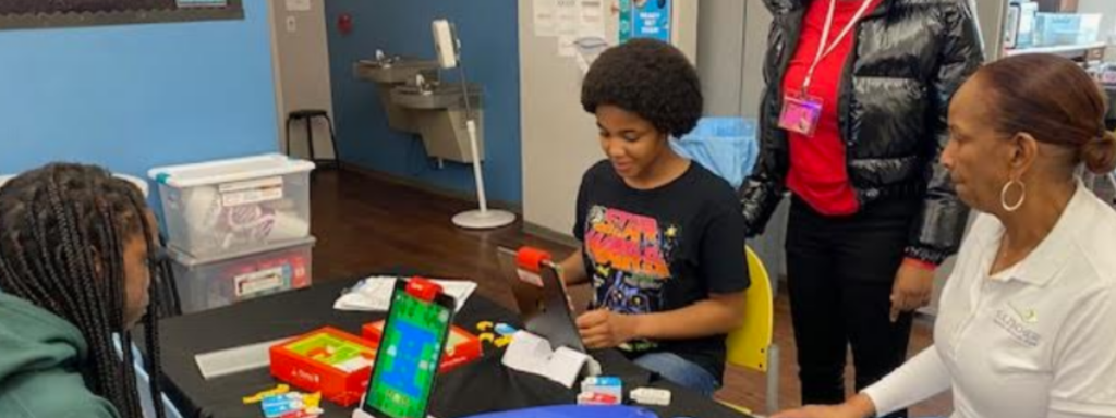 Young people working with educational robotics or coding materials at a table, with colorful electronic components and a tablet display visible.