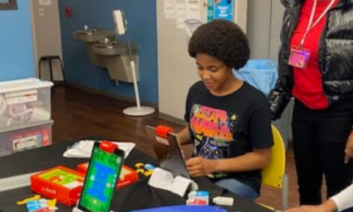 Young people working with educational robotics or coding materials at a table, with colorful electronic components and a tablet display visible.
