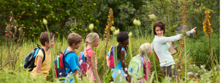 A teacher guides students through an outdoor learning experience, pointing out features in a garden or natural area. Students wearing colorful backpacks follow her lead.