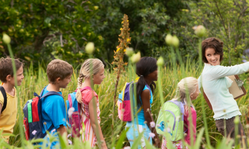 A teacher guides students through an outdoor learning experience, pointing out features in a garden or natural area. Students wearing colorful backpacks follow her lead.